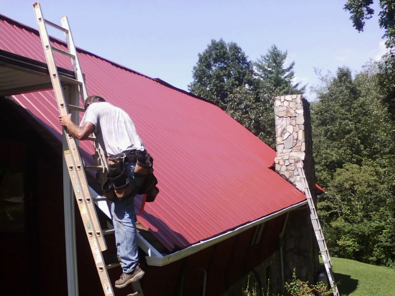 Roofer on a ladder beside a red metal roof and stone chimney — Soffit & Fascia Repair by Roofing Brothers in Malcom